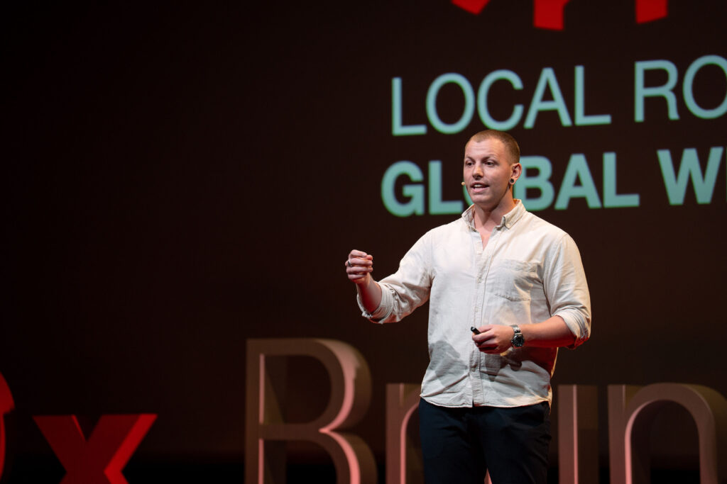 Ein Speaker im weißen Hemd spricht vor dem LOCAL ROOTS GLOBAL WINGS-Motiv