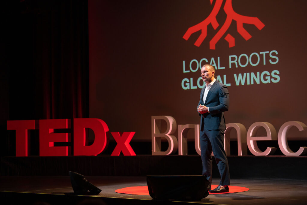Ein Speaker im blauen Sakko gestikuliert vor dem LOCAL ROOTS GLOBAL WINGS-Motiv